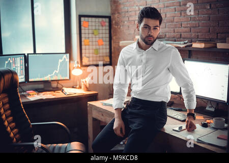 Successful attractive businessman or stock trader broker looking at camera posing at office desk with exchange graphs on computer monitors, young cryp