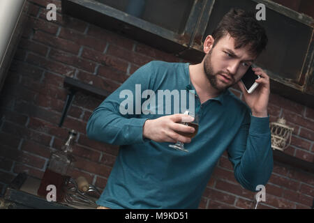 Young man alcohol addiction problems standing using smartphone talking serious conversation drinking whiskey Stock Photo