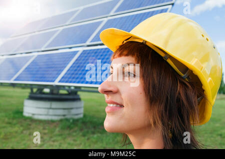 Side head portrait of a young woman with yellow construction site helmet in front of photovoltaic panel looking contented and smiling straight ahead. Stock Photo