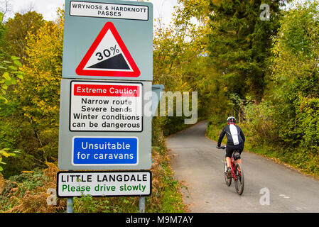 Cyclist passes warning signs on the approach to Wrynose and Hardknott passes in Little Langdale, the Lake District Stock Photo