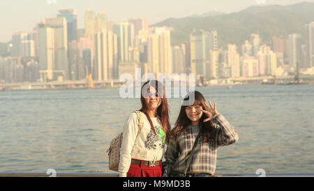 Hong Kong, China - January 1, 2016: Two young chinese tourist girls posing positively on the coast in Hong Kong, against a backdrop of a city panorama from Victoria harbor. Stock Photo