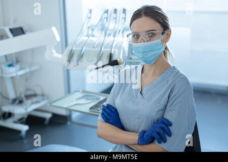 Ready for treatment. Pretty young female dentist wearing safety goggles, face mask and rubber gloves while posing for the camera and folding her hands Stock Photo