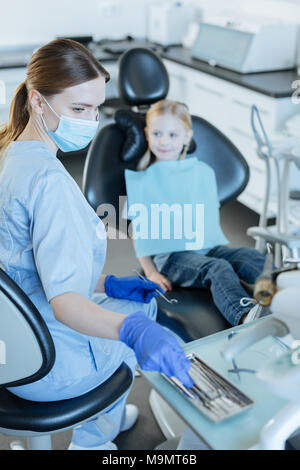 Thorough examination. Charming young female dentist conducting an examination of her little patient and picking a necessary instrument from the tray Stock Photo