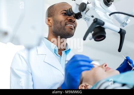 Examining in all details. Cheerful young male dentist using a professional microscope and conducting a checkup of his patients mouth cavity Stock Photo