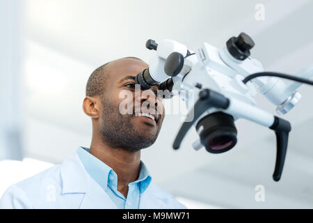 Advanced user. Pleasant cheerful male dentist smiling widely and looking into the lens of a microscope while examining his patients teeth Stock Photo