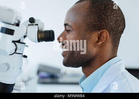 State-of-the-art device. The side view of a pleasant upbeat male dentist smiling widely while using a microscope at work and checking patients oral ca Stock Photo