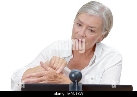 Elderly woman in front of a computer monitor with an attached webcam showing her doctor a mole on her hand during video call, isolated on white. Stock Photo