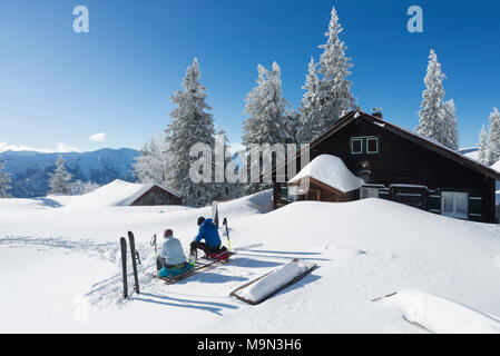 Man and woman with touring skis sitting on a bench in the snow at the Hubertus hut on the Mount Breitenstein in the brilliant winter sun, Bavaria Stock Photo