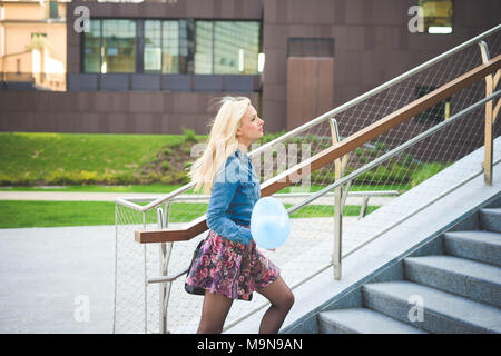 young beautiful blonde girl walking outdoor in the city park overlooking on her right wearing a jeans shirt, a hat and a floral skirt with a baloon in Stock Photo