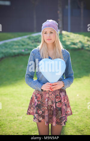 Knee figure of a young beautiful blonde girl posing outdoor in the city park overlooking on her right wearing a jeans shirt, a hat and a floral skirt  Stock Photo