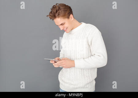 Young guy wearing stylish white sweater studio isolated on grey wall using smartphone joyful playing online games Stock Photo