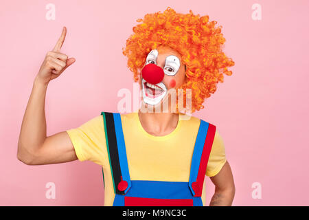 Man in a red wig, overalls and makeup finger up. Clown, corcus or show concept. Studio shot, pink background Stock Photo
