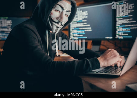 Masked hacker sit in workplace in corporate data center . Indoor, studio shot. Hacker attack, virus, net security concept Stock Photo