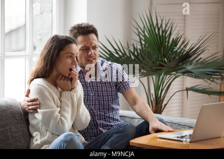 Scared man embracing shocked woman watching thrilling horror film or scary movie on laptop together, young couple feeling frightened and confused look Stock Photo
