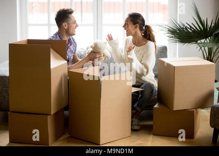 Young happy couple having fun packing boxes on moving day, loving boyfriend making gift holding cute toy found in belongings giving white teddy bear t Stock Photo