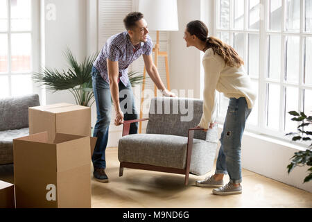Smiling couple carrying modern chair together placing furniture moving into new home, young family discussing house improvement interior design while Stock Photo