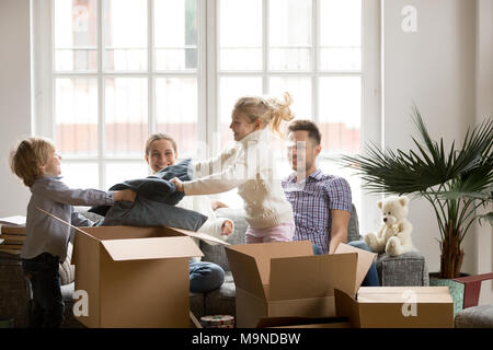 Happy children having fun pillow fight while family packing boxes together on moving day, parents laughing looking at kids playing on sofa in new hous Stock Photo