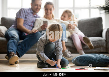 Serious cute son holding color marker drawing on floor while parents with sister sitting apart, preschool kid loner boy playing alone, family leisure Stock Photo