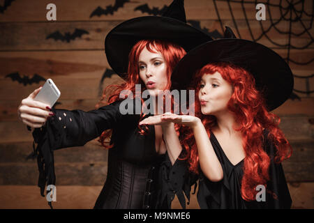 Halloween Concept - Beautiful caucasian mother and her daughter with long red hair in witch costumes taking a selfie with smartphone  ready for celebrating Halloween. Stock Photo