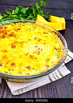 Pumpkin kish with pumpkin and bacon, filled with milk and eggs in a glass frying pan on a napkin, parsley, vegetable slices against a dark wooden boar Stock Photo