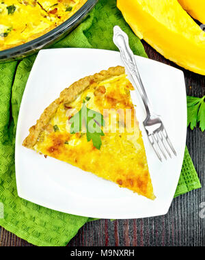 Piece of pie kish with pumpkin and bacon, filled with milk with eggs and cheese in a plate on a napkin, parsley on a wooden board background from abov Stock Photo