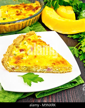 Piece of pie kish with pumpkin and bacon, filled with milk with eggs and cheese in a plate on a green napkin, parsley on a background of a dark wooden Stock Photo