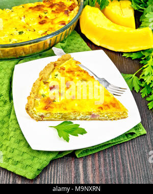 Piece of pie kish with pumpkin and bacon, filled with milk with eggs and cheese in a plate on a napkin, parsley on a wooden board background Stock Photo