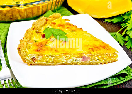 Piece of pie kish with pumpkin and bacon, filled with milk with eggs and cheese in a plate on a napkin, parsley on a background of a dark wooden board Stock Photo