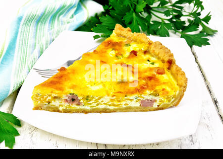 Piece of pie kish with pumpkin and bacon, filled with milk with eggs and cheese in a plate, parsley, a towel on the background of a light wooden board Stock Photo