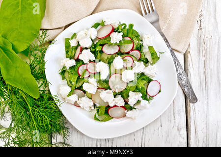 Salad with spinach, cucumbers, radish and salted cheese, dill and green onions in a bowl, towel on the background of a wooden board from above Stock Photo