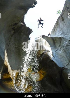 A man in wetsuit jumping from a high cliff next to a waterfall during a canyoning adventure trip in Switzerland. Ticino, in the southern part of Switzerland is a small paradise for canyoning thanks to numerous little waterfalls, torrents and rivers that rush down the valley through the wilderness. Cresciano, Ticino Canton, Switzerland Stock Photo