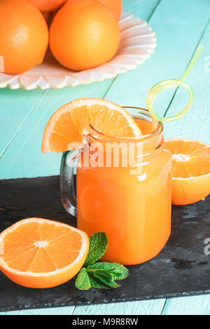 Mason jar full of fresh orange juice, with a funny straw, surrounded by sliced oranges and mint leaves, and a plate of oranges in thr background. Stock Photo