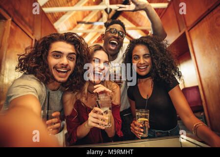 Group of happy young friends with drinks gathered for party in club. Excited men and woman with drinks taking selfie during party. Stock Photo