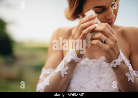 Beautiful bride in white dress weeps tears of happiness on the wedding day. Emotional woman in wedding gown wipes the tears with tissue paper. Stock Photo