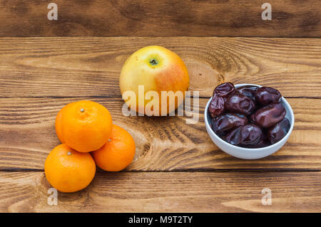 Four bright orange ripe mandarins, an apple and a cup with dates on a wooden table. Stock Photo