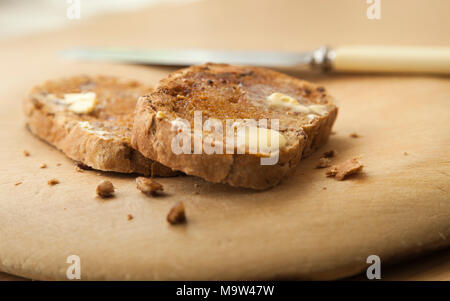 Two slices of warm nutty artisan bread toasted with melting butter on wooden board with a knife out of focus beside the slices. Stock Photo