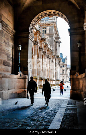 Three people walking with briefcases and bags two advancing through passageway arch of the Louvre, as silhouettes casting shadows; a sense of scale Stock Photo