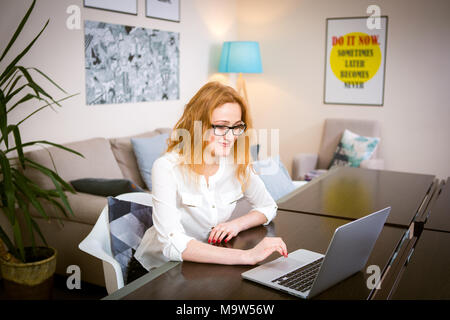 Young woman with long red hair and glasses for view works, prints on a gray laptop keyboard sitting at a wooden table in a bright interior. Subject bu Stock Photo