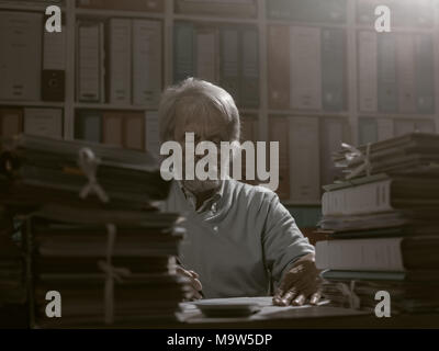 Overloaded senior office worker surrounded by stacks of papework, he is writing on a document Stock Photo