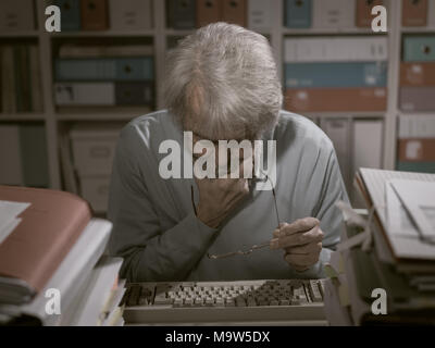 Senior office worker using an obsolete keyboard, he is having difficulties and thinking with hand on chin Stock Photo