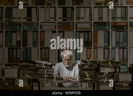 Frustrated senior office worker sitting at office desk and overloaded with work, his desk is covered with piles of paperwork Stock Photo