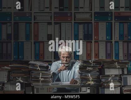 Senior office worker sitting and office desk overloaded with work and being lazy, he is balancing a pen on his finger Stock Photo
