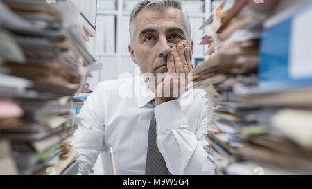 Frustrated overwhelmed executive working in the office and overloaded with paperwork, he is leaning on his arm and feeling depressed Stock Photo