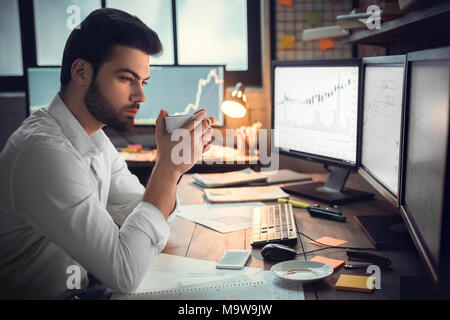 Thoughtful serious trader broker drinking coffee working overtime looking at computer monitors analyzing stock trading market graphs, overwork, lack o Stock Photo