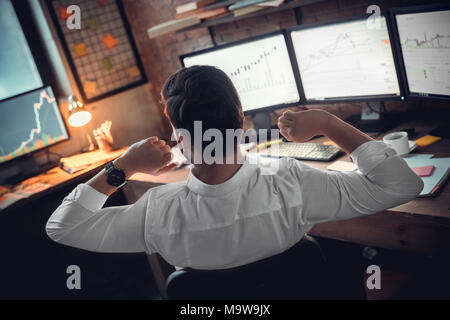 Rear view at stock trader broker or investor stretching hands at workplace tired after long using computer, businessman finished work taking break for Stock Photo
