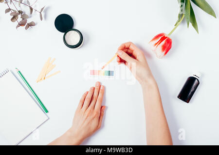 Woman testing cosmetics by using litmus paper and scale. Top view of female hands measuring of pH level in cosmetics among tulip, eucalyptus branch an Stock Photo