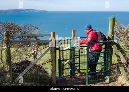 Elderly active senior woman walker walking through a new metal kissing gate near coast with sea beyond. Llanddona Isle of Anglesey Wales UK Britain Stock Photo