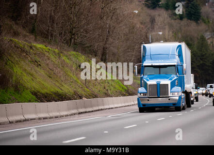 Modern blue powerful stylish big rig tall cab semi truck fleet for long haul deliveries with semi trailer going on wide highway with trees on the hill Stock Photo