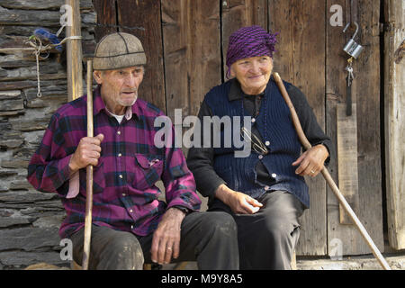 An elderly couple holding walking sticks rest on a bench outside an old stone shed, Ushguli, Upper Svaneti, Georgia Stock Photo