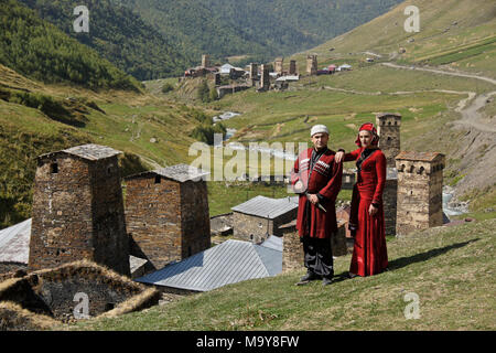 A man and woman in Georgian national dress stand on a hill above stone buildings and medieval defensive towers in Ushguli, Upper Svaneti, Georgia Stock Photo